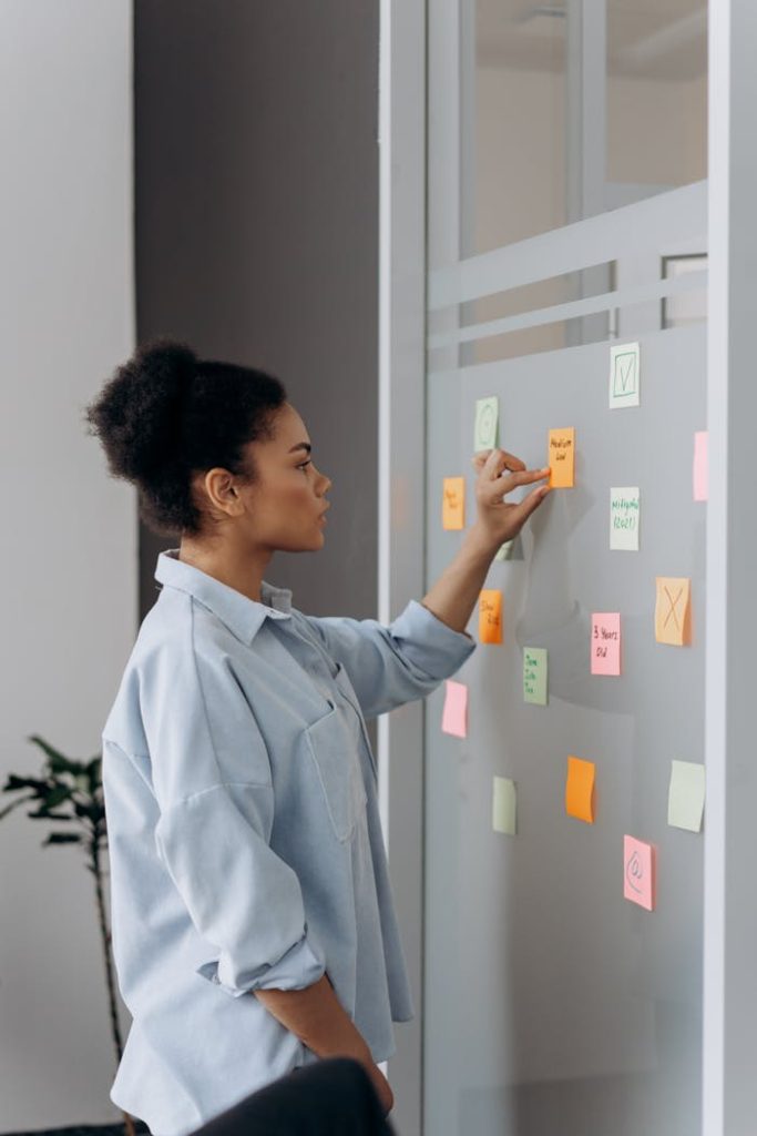 Young professional organizing tasks on a glass board with colorful sticky notes in an office setting.